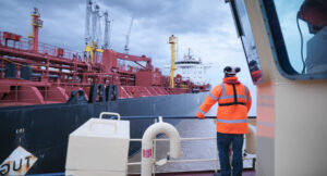 Worker in an orange jacket observes a large red industrial ship at port