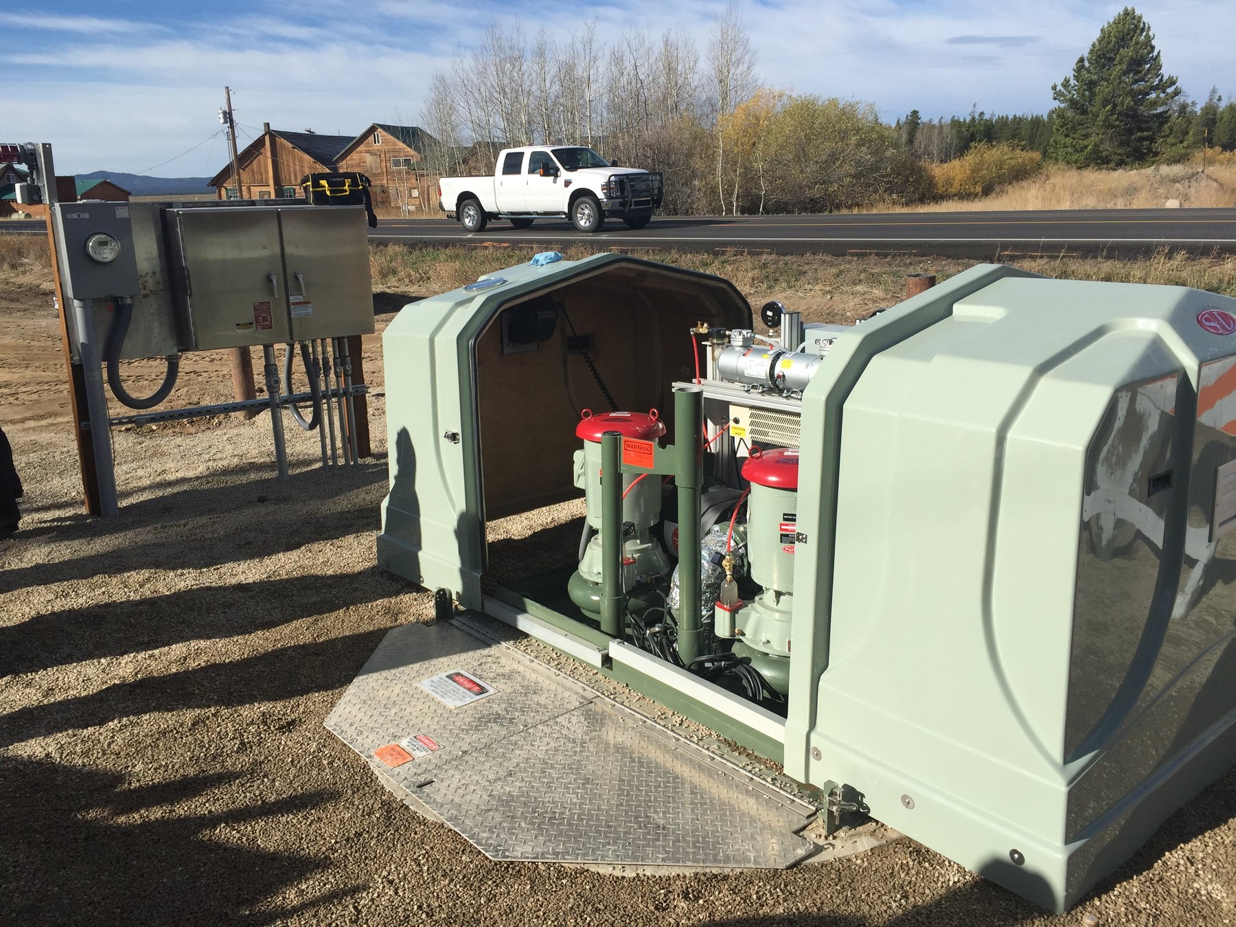 Packaged above-ground pump station with open sliding enclosure beside a road, with control cabinet on the left