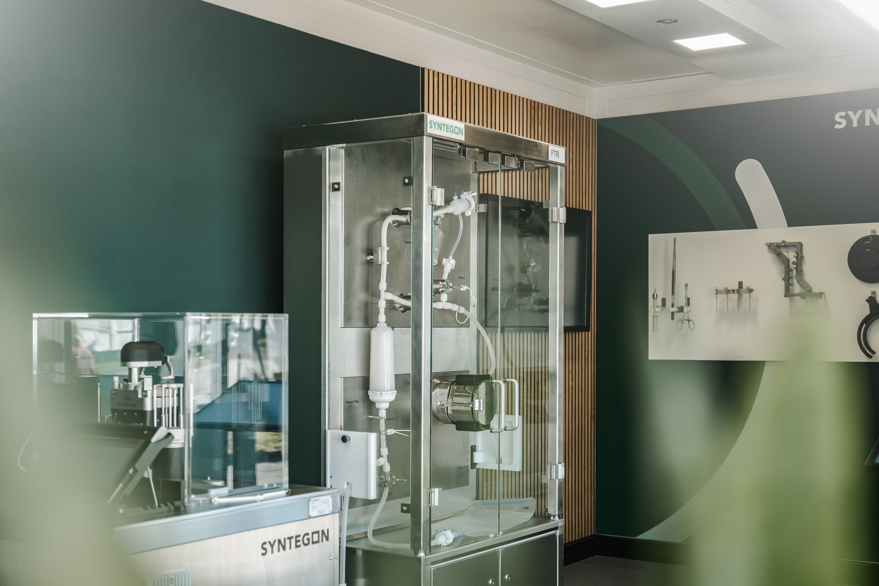 Exhibits inside the Syntegon roadshow trailer, including a PUPSIT filter test rack in a glass enclosure and a settle plate changer