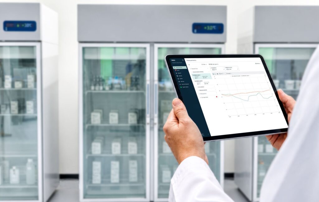 A lab professional holding a tablet showing the Vaisala Viewlinc Cloud Suite measurements screen in front of laboratory refrigerators