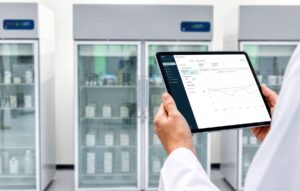A lab professional holding a tablet showing the Vaisala Viewlinc Cloud Suite measurements screen in front of laboratory refrigerators